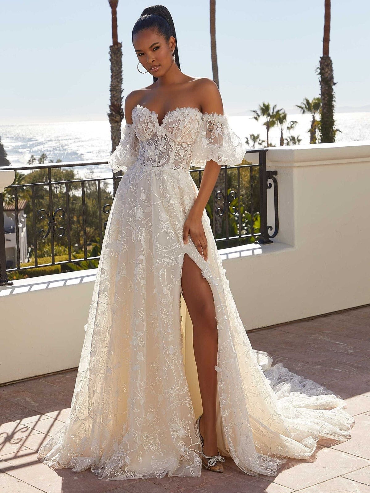 Bride in the Morilee Magdalena gown, an off-the-shoulder lace A-line with puff sleeves, a front slit and a train, on a seaside terrace.